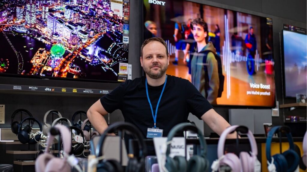 Člen týmu Sales House Group stojí v prodejně elektroniky a představuje práci v promo oddělení se zaměřením na zákazníky a produkty.