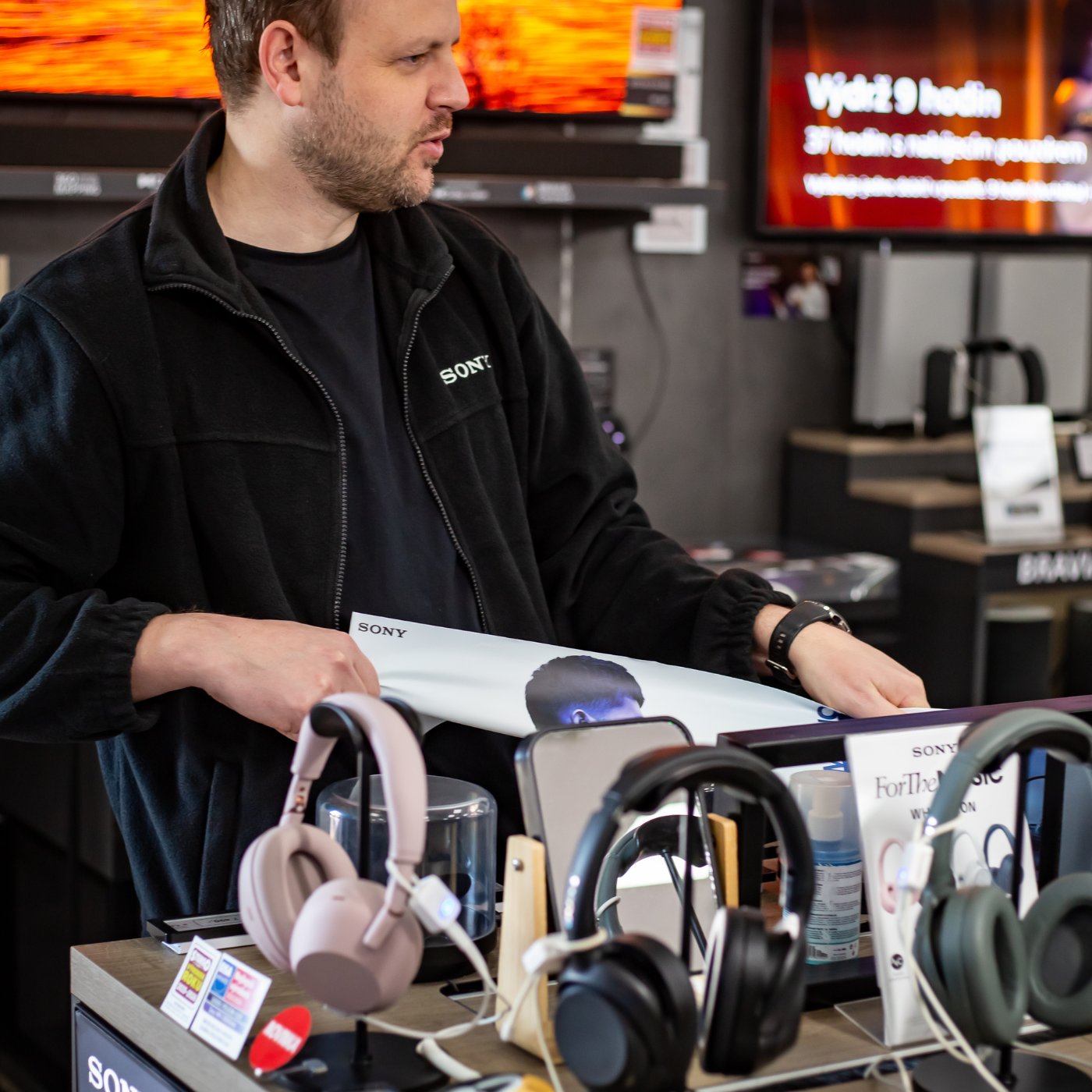 Promotér Sales House Group v černé bundě Sony připravuje reklamní banner na stojanu s prémiovými sluchátky v prodejně elektra.