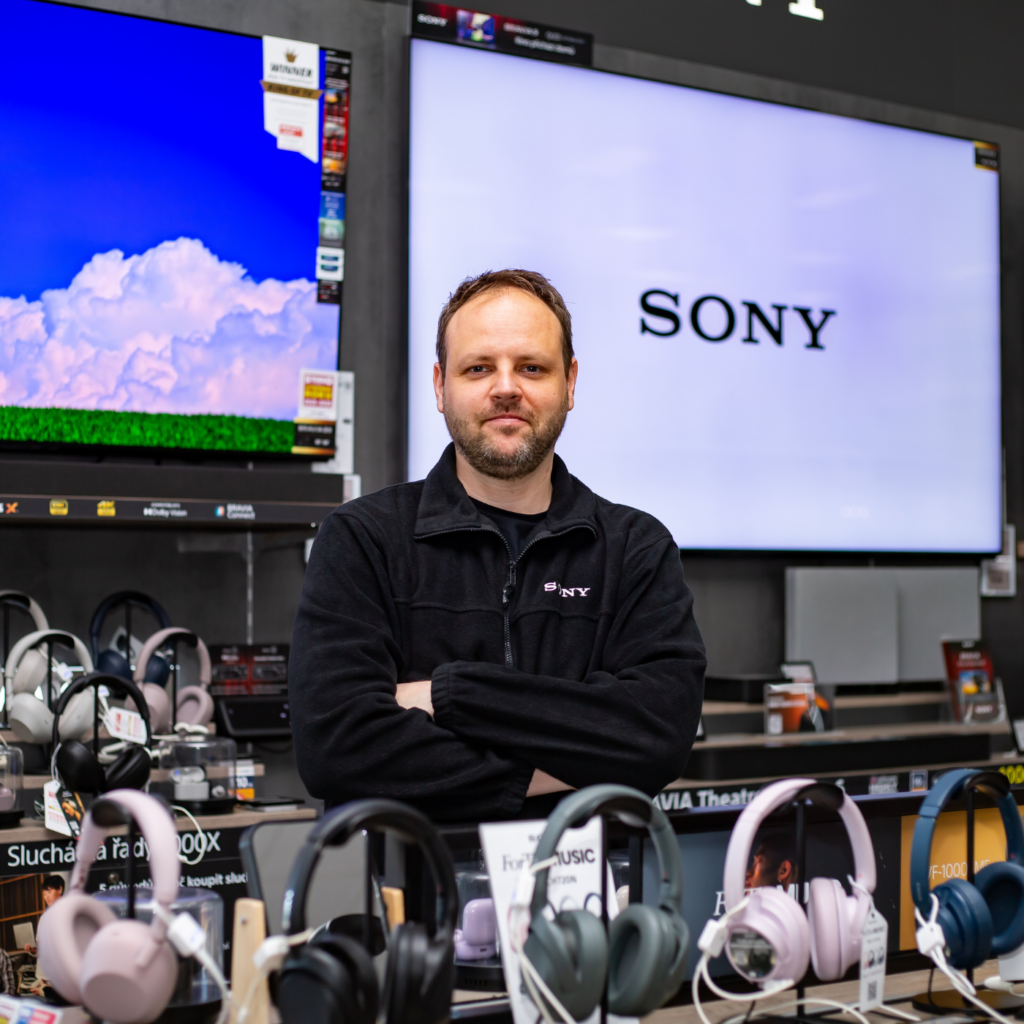 Usměvavý promotér Sales House Group v černé mikině Sony stojí se založenýma rukama u vystavených sluchátek a televizí v prodejně elektra.