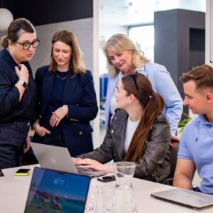 Pětičlenný tým zaměstnanců Sales House Group v moderní kanceláři – kolegové stojí a sedí kolem stolu a společně diskutují nad prací na notebooku.